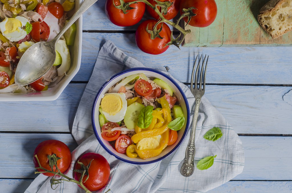 su tavolo di legno bianco, ciotola con gli ingredienti dell'insalata nizzarda e pomodori accanto