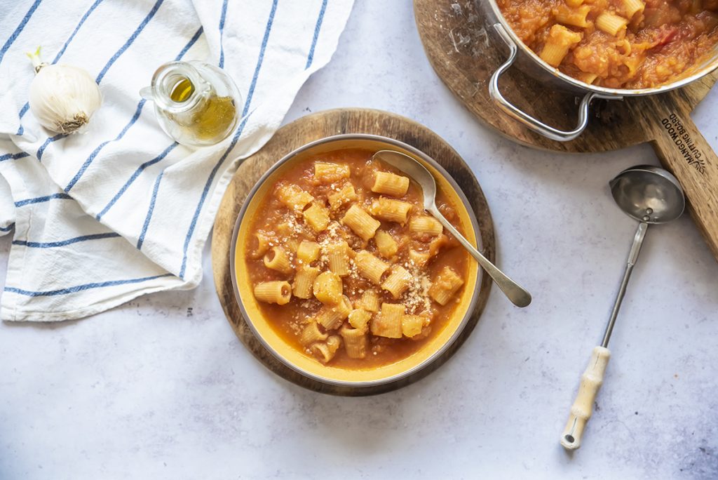 su piano da cucina pasta e patate, pentola con pasta e patate, un piatto pronto con cucchiaio, 1 mestolo, 1 canovaccio, aglio e 1 piccolo dosatore di olio EVO