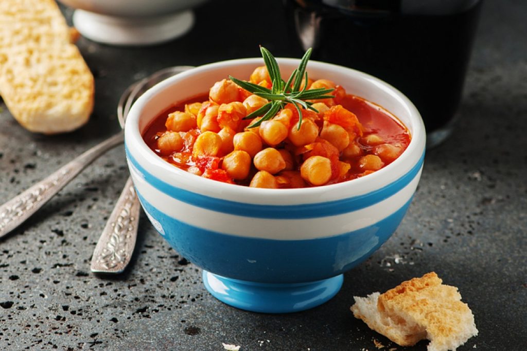 su tavolo nero un ciottola blu e bianca con una zuppa di ceci e rosmarino con accanto dei crostini e dietro 2 cucchiai