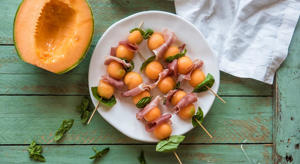 su tavolo di legno verde, piatto bianco con spiedini di prosciutto e melone e basilico con accanto melone tagliato e canovaccio bianco