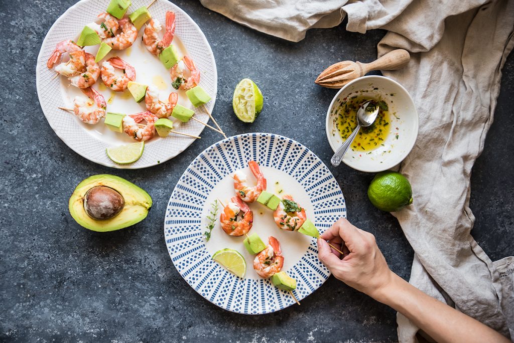 su tavolo grigio piatto di spiedini di gamberi con avocado. Una mano che li adagia sul piatto, accanto avocado tagliato in 2 e la marinatura in una ciotola