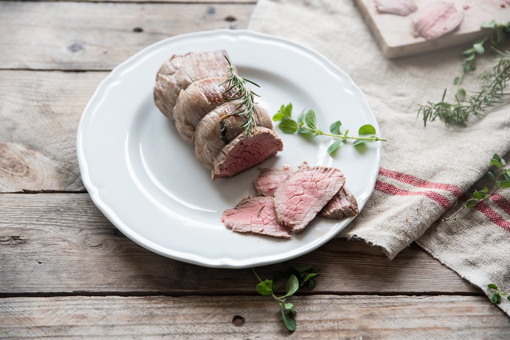 su tavolo di legno piatto bianco con un roast beef e alcune fette tagliate accanto erbe aromatiche