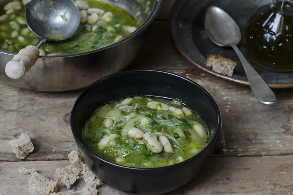 Su tavolo di legno, pentola con zuppa di scarola e fagioli con un mestolo. Accanto piatto di zuppa pronta con cucchiaio