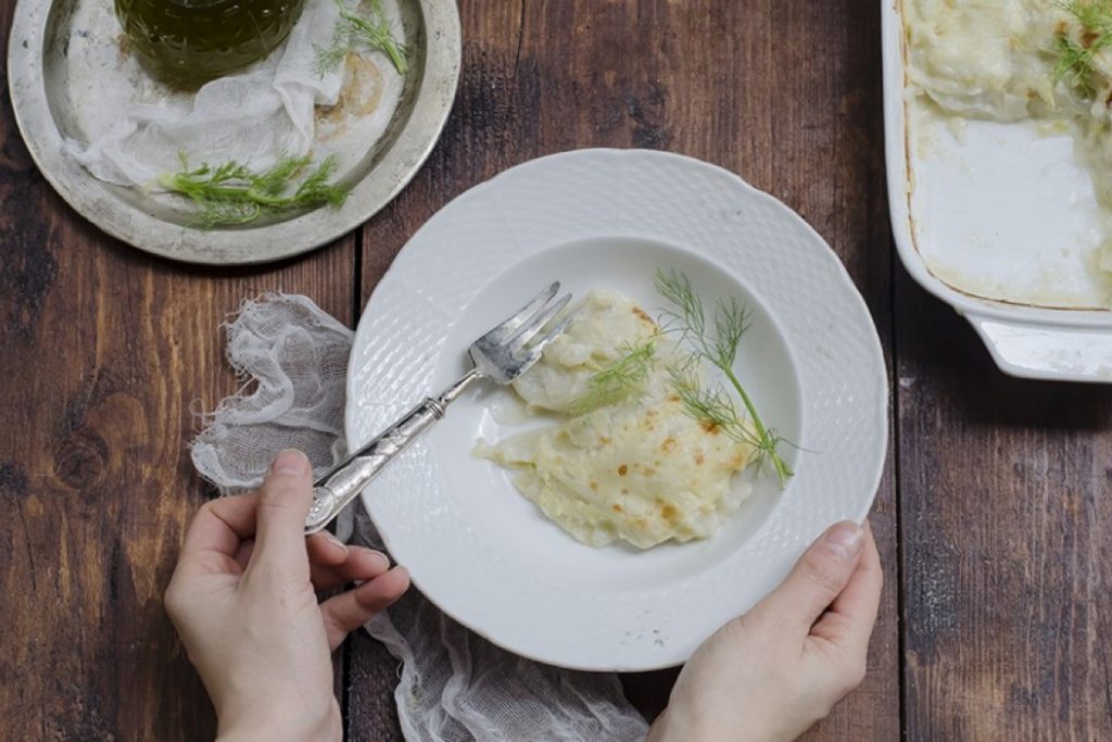 mani tengono una scodella bianca con finocchi gratinati, forchetta