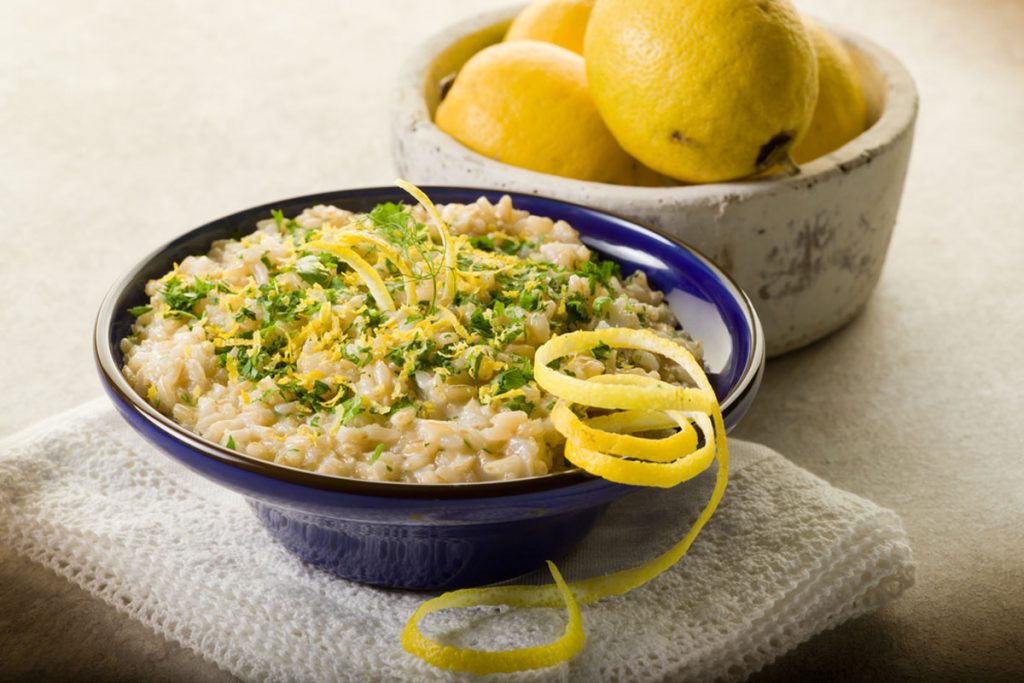 su sfondo chiaro ciotola blu con un risotto al limone e dietro ciotola con limoni biologici