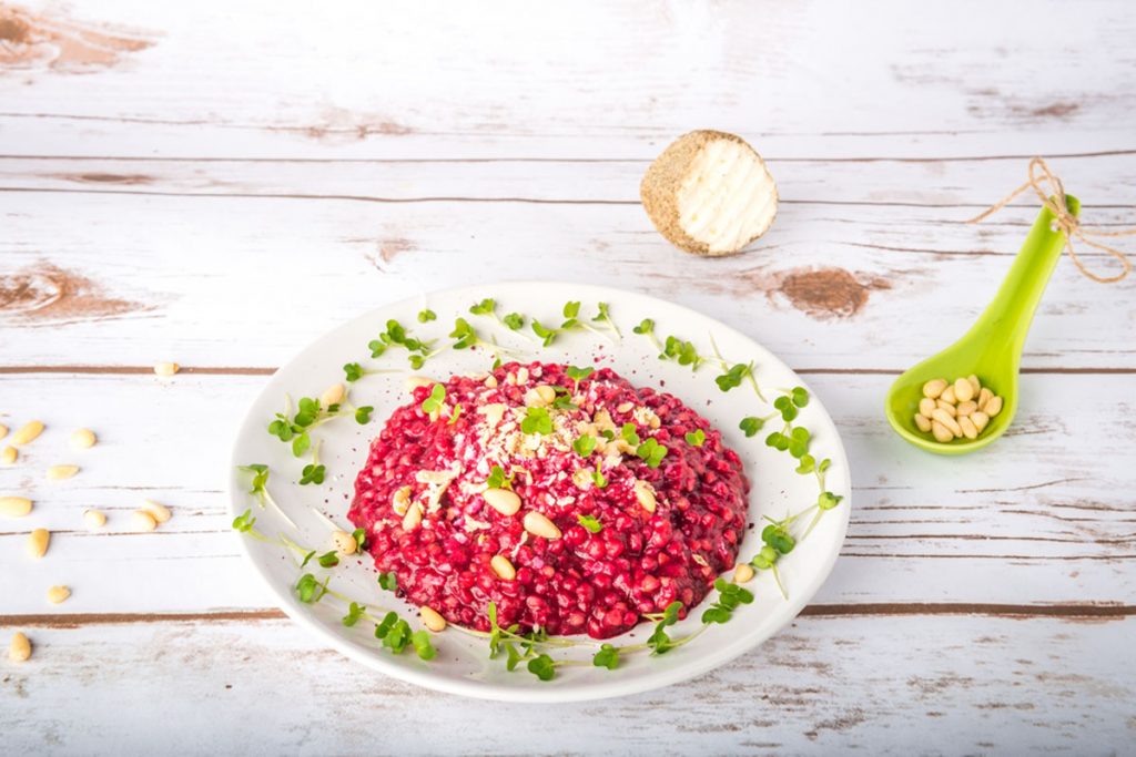 su tavolo di legno bianco un piatto bianco con un risotto alla barbabietola rossa decorato con semi e frutta secca. Accanto cucchiaio verde con pinoli