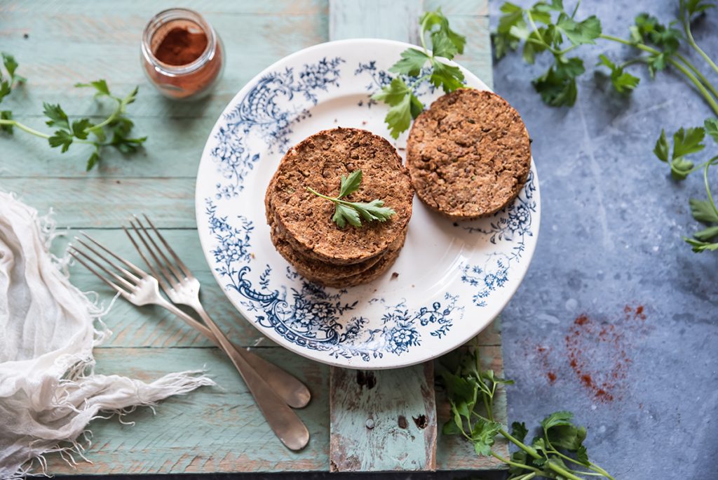 su tavolo di legno azzurro, erbe aromatiche sparse con piatto bianche e blu con 2 burger di lenticchie a destra del piatto 2 forchette e dietro contenitore con paprika