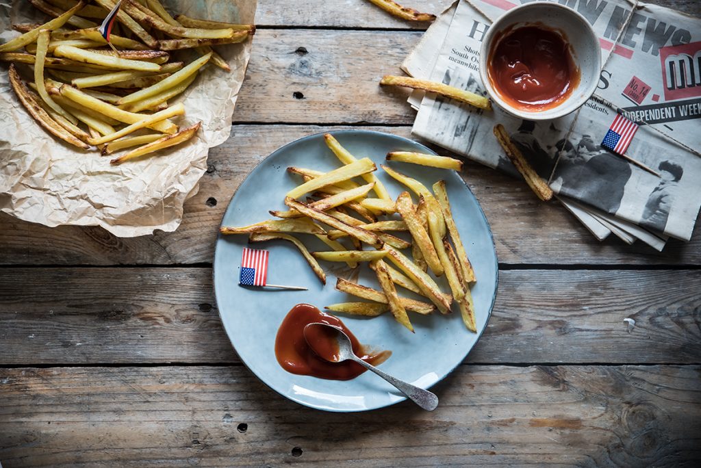 su tavolo di legno, piatto azzurro con patatine fritte al forno e un cucchiaino di ketchup e una bandierina degli USA. Dietro, giornali cin un contenitore di ketchup e sulla destra carta da forno con altre patatine