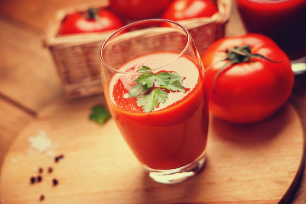 su un tagliere di legno, un bicchiere con succo di pomodoro e prezzemolo, con accanto un pomodoro San Marzano e pepe nero