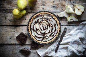 teglia con crostata pere e cioccolato, accanto a pere intere, pere tagliate, pezzi cioccolato, un coltello e un canovaccio