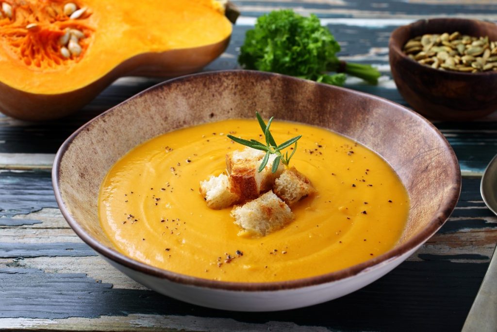 zuppiera rotonda con vellutata di zucca e alcuni crostini al centro, mezza zucca sullo sfondo