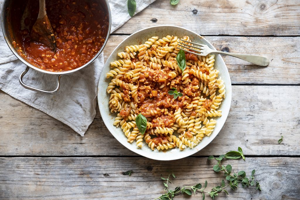 su base di legno un piatto chiaro pieno di fusilli con ragù di tempeh, accanto un pentolino con salsa di ragù