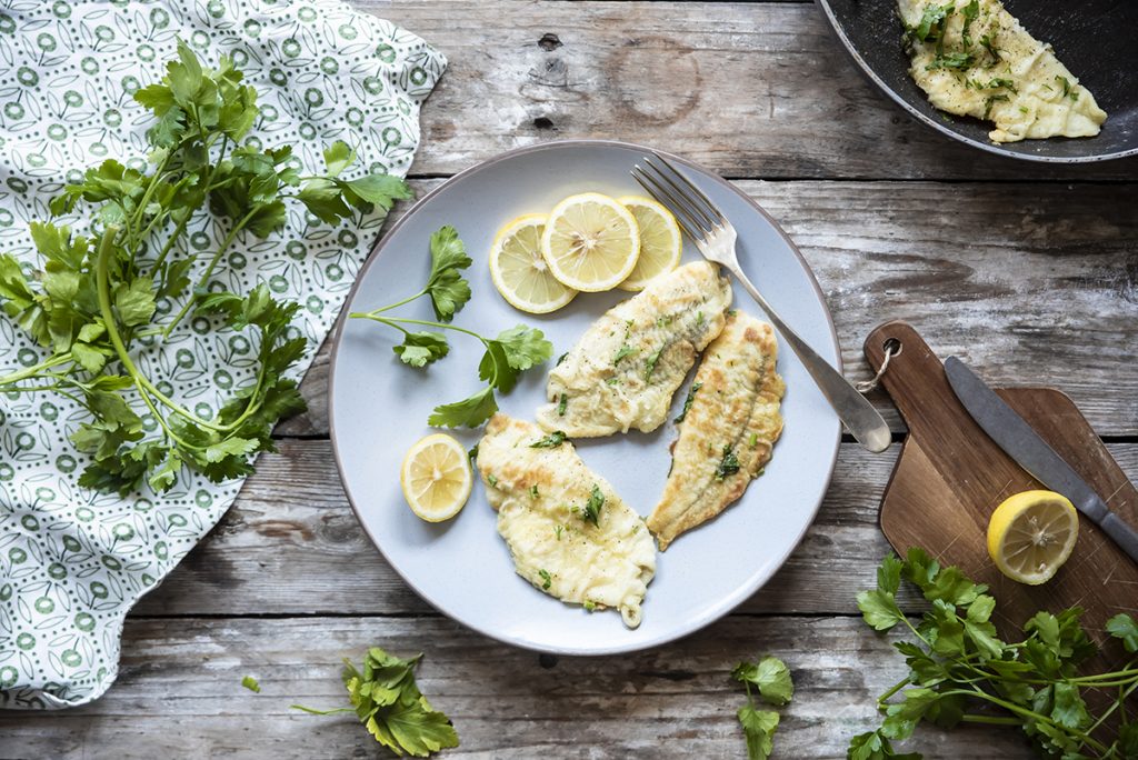 su tavolo di legno un piatto bianco con platessa alla mugnaia, erbe aromatiche attorno e una padella con un altra fetta di platessa