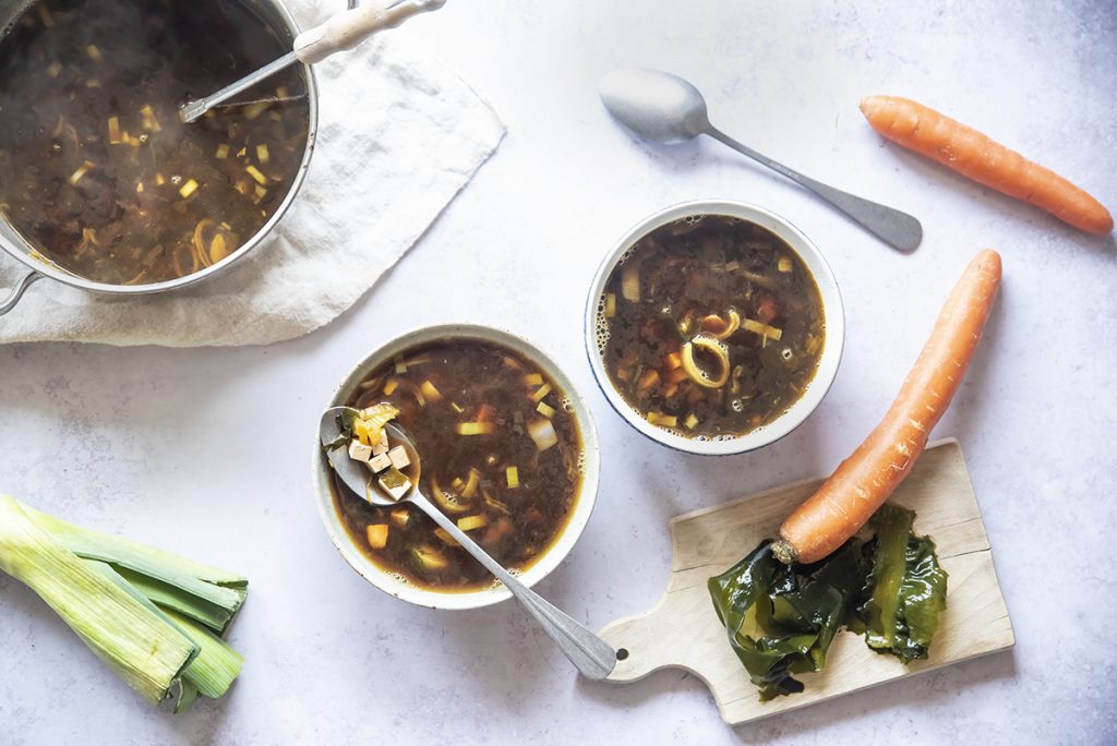 su tavolo di marmo 2 contenitori bianchi con la zuppa di miso e attorno gli ingredienti: carota ,1 porro 2 cucchiai, e dietro la zuppiera piena di zuppa