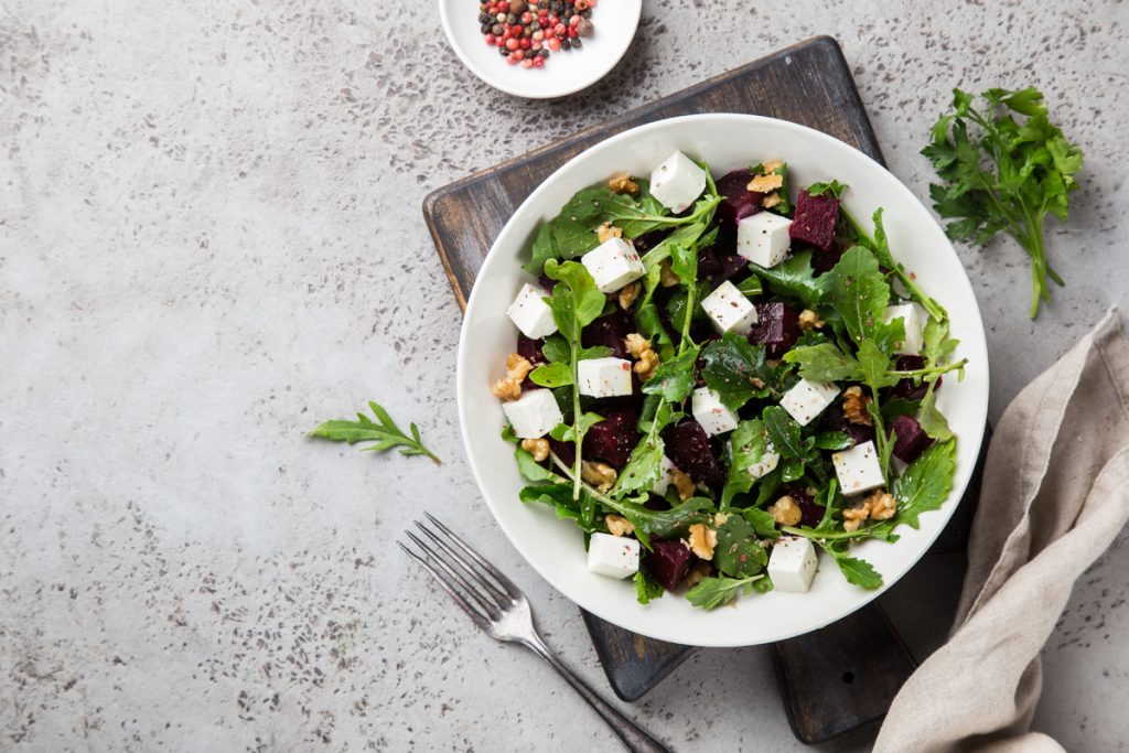 su piano di marmo con sotto tagliere nero piatto bianco con rucola, fetta e frutta secca. Accanto peperoncino e forchetta vicino al piatto