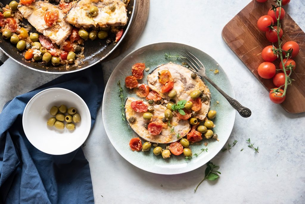 su tavolo bianco di marmo piatto verde con pesce alla siciliana e ciotola con olive verdi e pomodorini su tagliere di legno