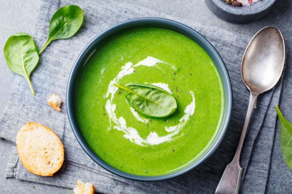 su canovaccio grigio, ciotola con vellutata di spinaci con accanto un cucchiaio, spinaci freschi e una bruschetta