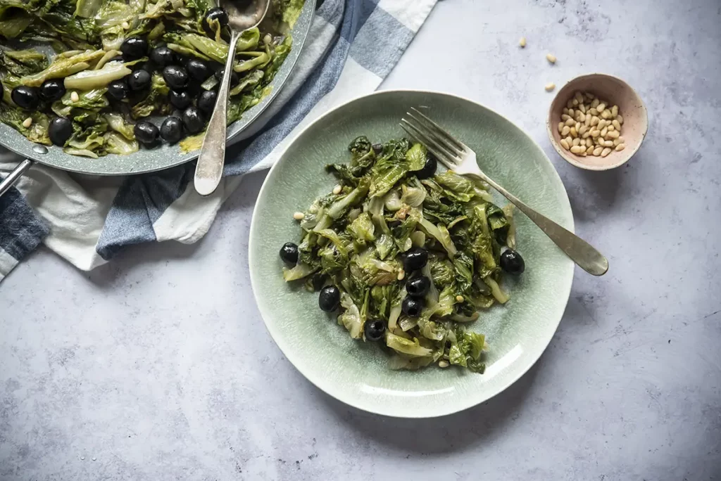 su piano di marmo, piatto verde con della scarola stufata con pinoli, accanto un contenitore piccolo con dei pinoli e dietro un canovaccio con un piatto di scarola