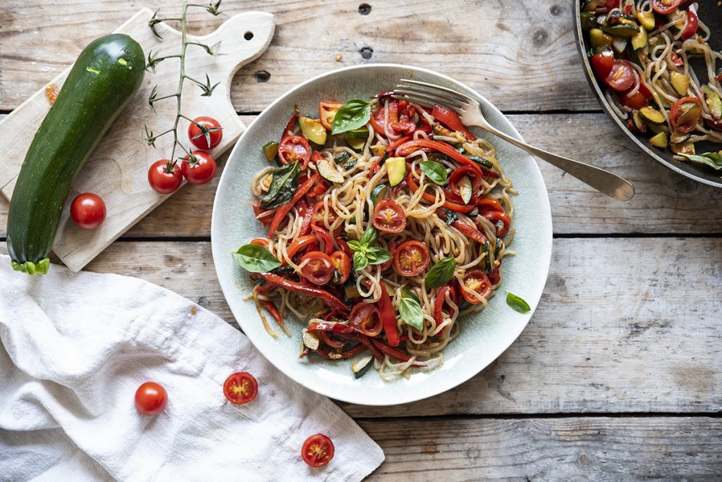 su tavolo di legno piatto con shirataki di konjac con verdure, accanto canovaccio con pomodorini e sul tagliere di legno zucchine . A destra padella con spaghetti di konjac