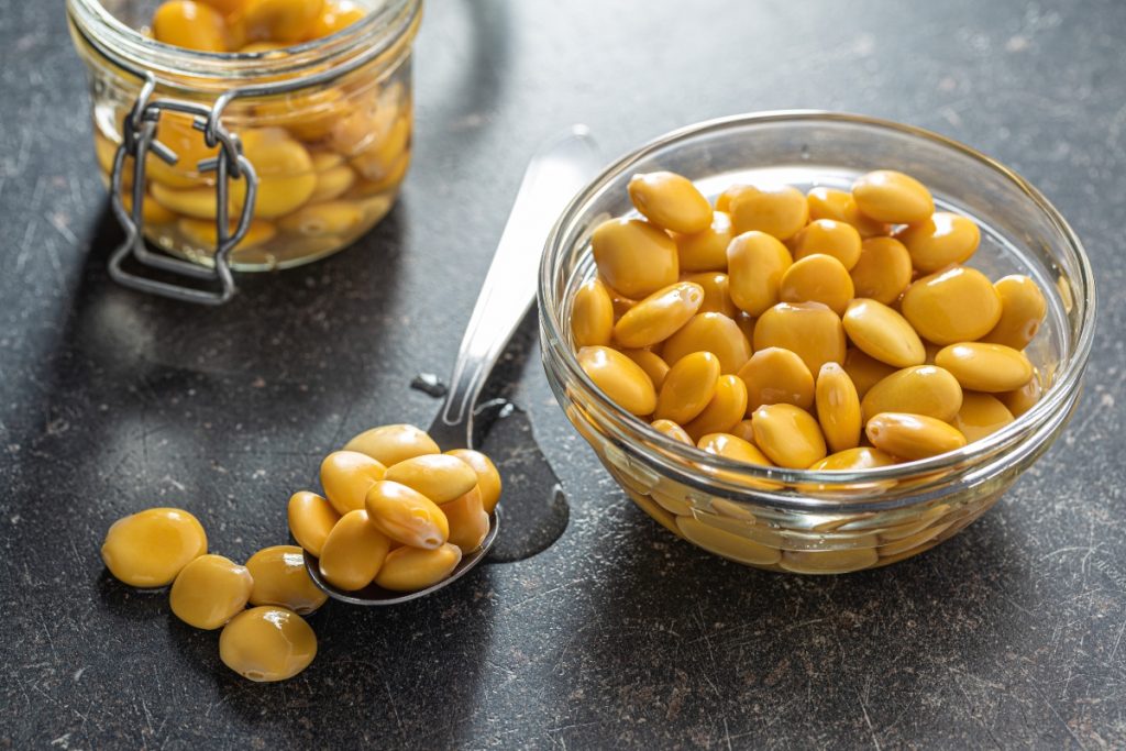 su tavolo nero, un contenitore di vetro pieno di lupini, un cucchiaio di lupini e dietro un barratolo pieno di semi di lupino