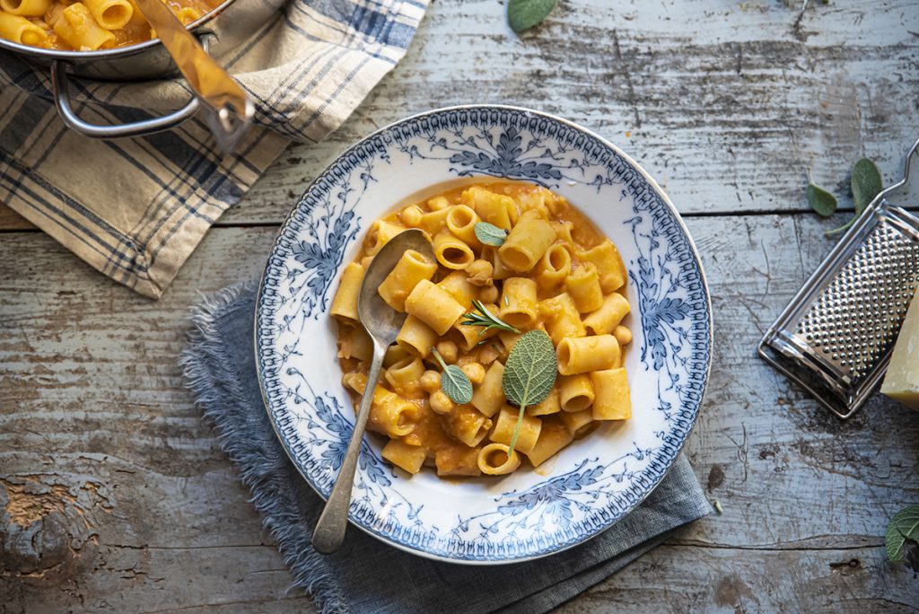 Su tavolo di legno piatto colorato con pasta e ceci e un foglio di alloro, accanto grattugia e il parmigiano. In alto a sinistra la padelle con pasta e ceci