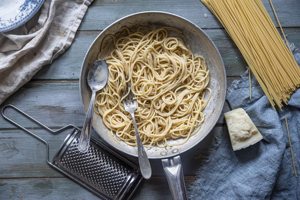 su tavolo di legno, in un padella spaghetti cacio e pepe, con accanto la grattugia, il Pecorino e gli spaghetti