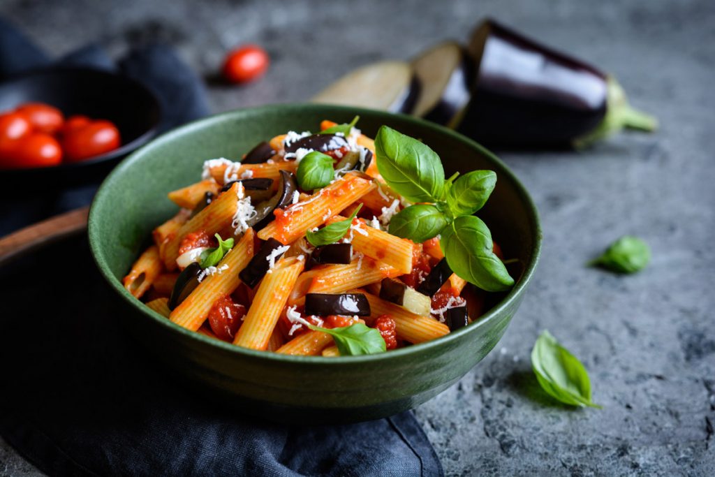 su tavolo di legno, ciotola verde con pasta alla Norma e attorno ingredienti, pomodori, melanzane e basilico