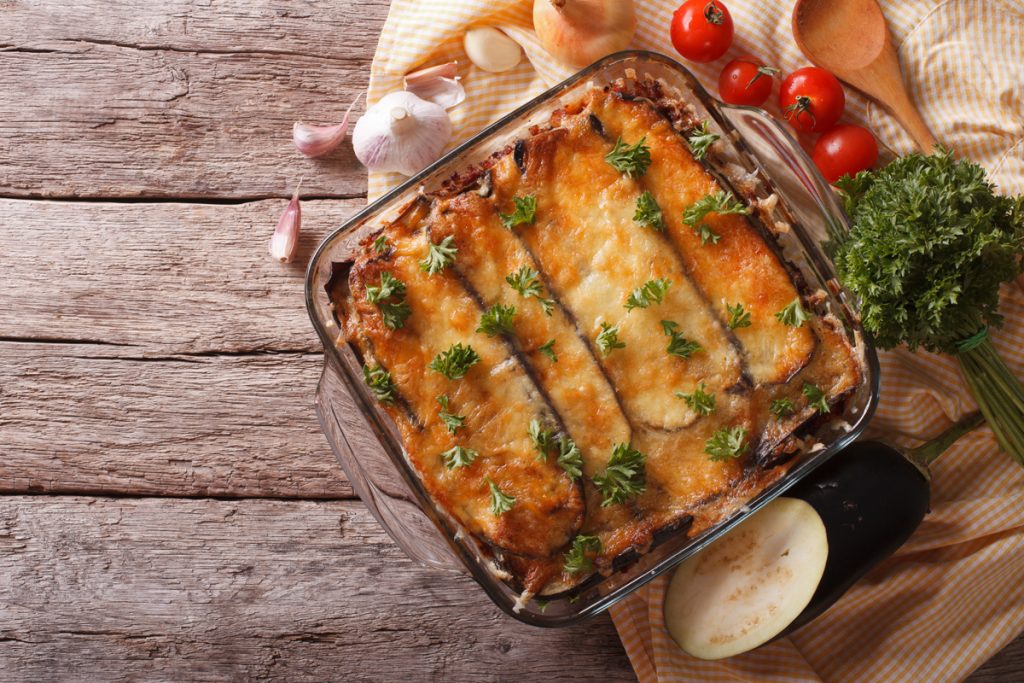 Su tavolo di legno, pirofila con la moussaka, aglio, pomodorini e erbe aromatiche