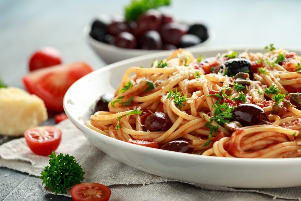 su tavolo primo piano di un piatto con spaghetti alla puttanesca, una ciotola con olive nere, aglio e pomodorini tagliati