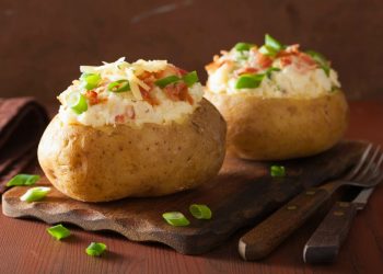 su tavolo di legno, tagliere di legno con 2 patate ripiene al forno, accanto forchetta e coltello