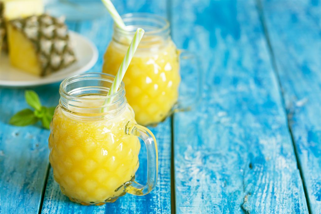 tavolo di legno azzurro con 2 bicchieroni pieni di succo di ananas e bromelina e cannucce e dietro un ananas tagliato