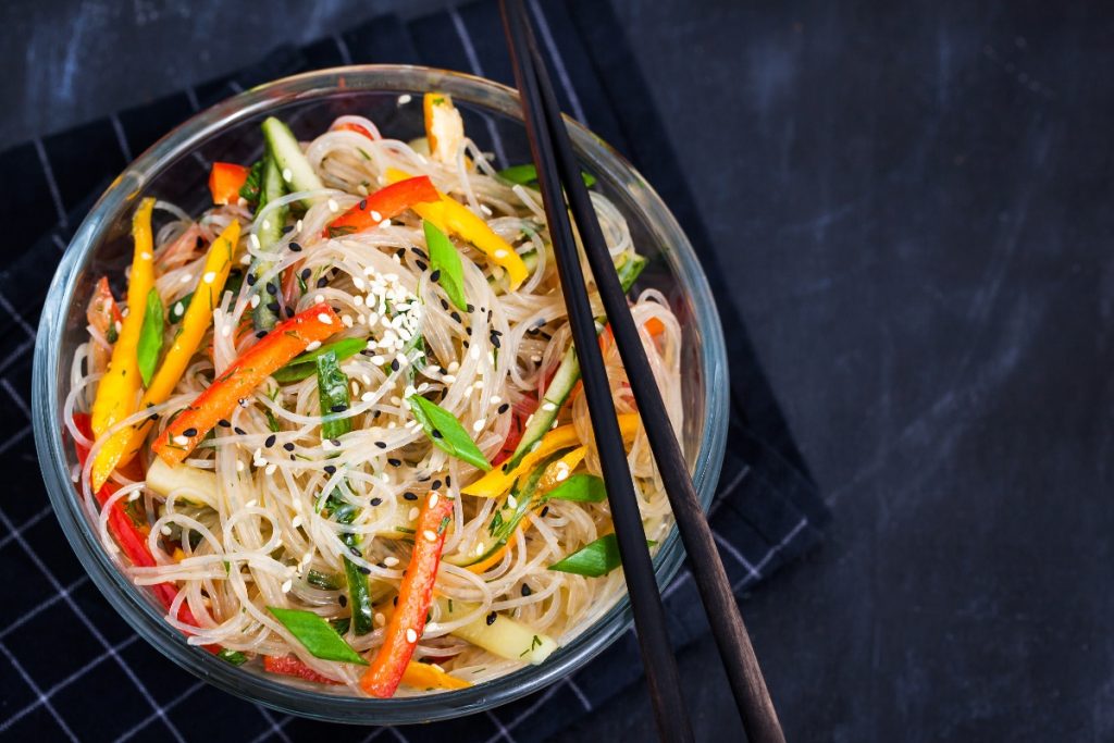 su sfondo nero ciotola nera con spaghetti di soia con verdure. peperoni rossi e verdi e carote. Sulla ciottola 2 bacchette cinesi per mangiarli