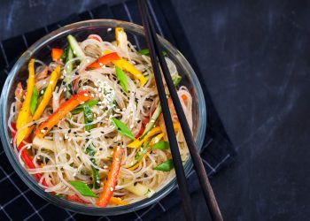 su sfondo nero ciotola nera con spaghetti di soia con verdure. peperoni rossi e verdi e carote. Sulla ciottola 2 bacchette cinesi per mangiarli