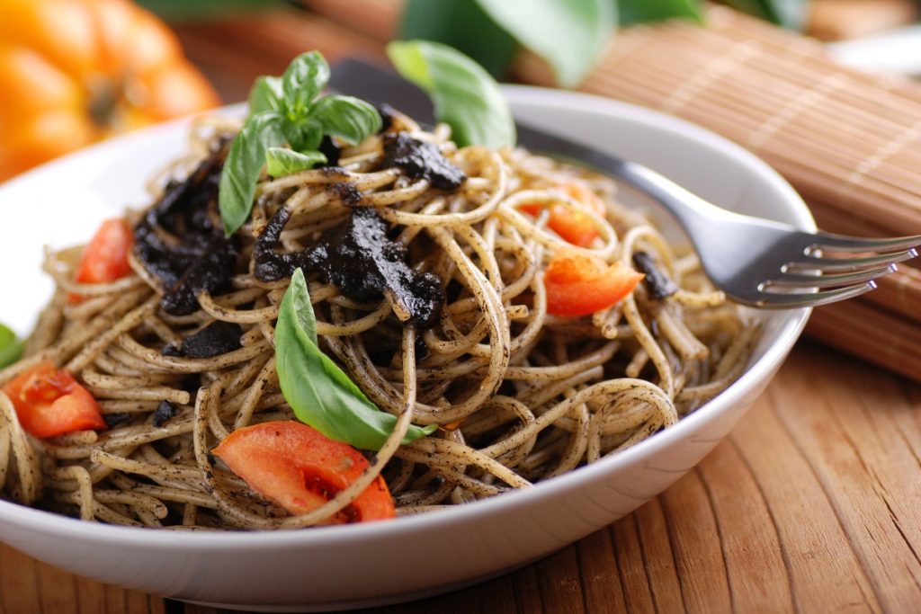 Primo piano di un piatto bianco con spaghetti al nero di seppia, pomodori e prezzemolo con una forchetta sul bordo. In secondo piano un pomodoro