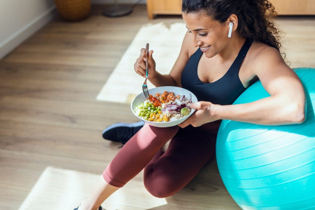 Donna in casa vestita da palestra, appoggiata su una fitball che mangia un insalata perché segue la dieta anticellulite