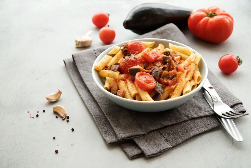 su tavolo bianco un tovagliolo grigio con sopra un piatto bianco con pasta con melanzane e pomodorini. A destra, forchetta e coltelli messi alla rovescia e dietro pomodori e melanzana