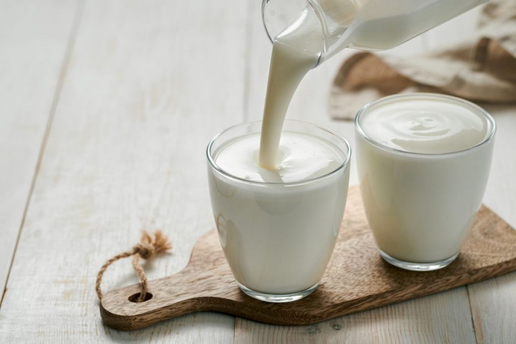 su tavolo di legno bianco, tagliere di legno con 2 bicchieri con yogurt fatto in casa e una mano che versa il latte sopra