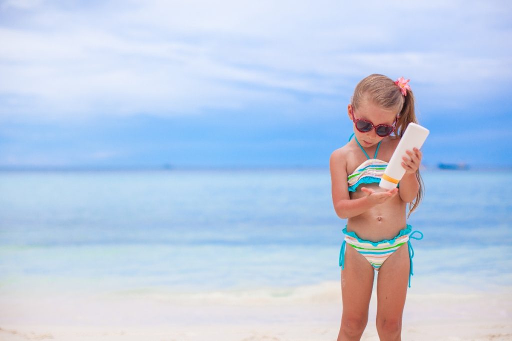 bambina sulla spiaggia con occhiali da sole e una crema solare in mano