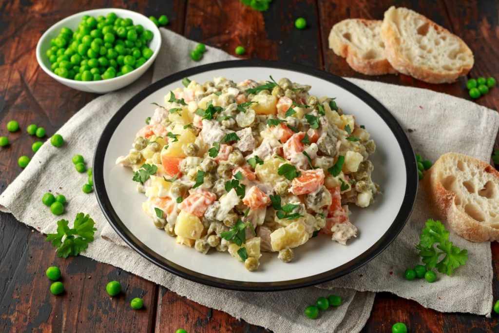 su tavolo di legno sotto piatto bianco con piatto bianco con insalata russa, accanto piselli e fette di pane