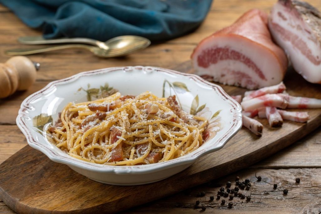 su tavolo di legno, tagliere di legno con un piatto di pasta alla Gricia e attorno del bacon tagliato, pepe nero e un cucchiaino