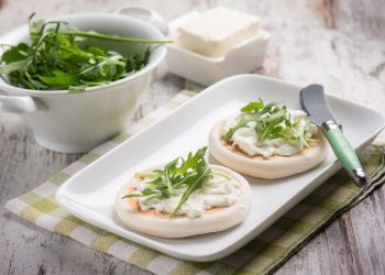 su tavolo di legno, tovagliolo a quadretti bianco e verdi con piatto bianco rettangolare e 2 tigelle di Modena con ricotta e stracchino . Accanto contenitore con rucola e in un'altro stracchino