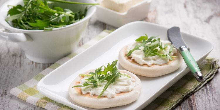 su tavolo di legno, tovagliolo a quadretti bianco e verdi con piatto bianco rettangolare e 2 tigelle di Modena con ricotta e stracchino . Accanto contenitore con rucola e in un'altro stracchino