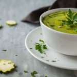 su base grigia piattino e tazza da brodo bianca piena di vellutata di zucchine. Rondelle di zucchine sparse