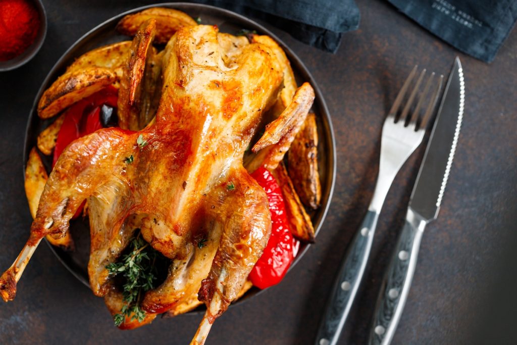 Primo piano di una piatto nero su tavolo da cucina con una faraona al forno, accanto forchetta e coltello