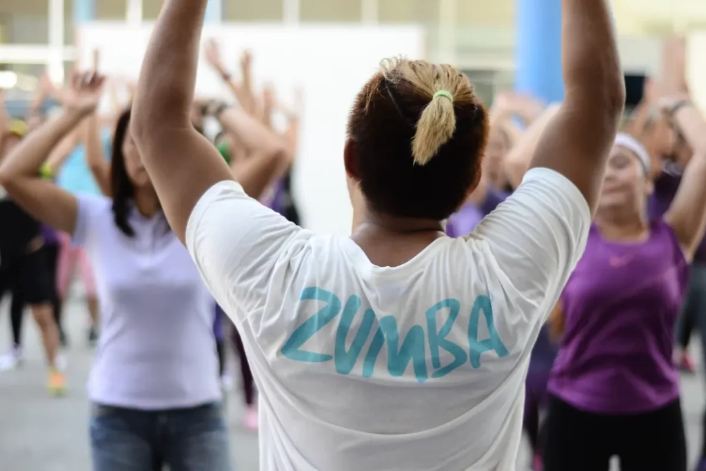 In una palestra, una lezione con un insegnante di schiena che indossa una maglietta bianca con la scritta zumba