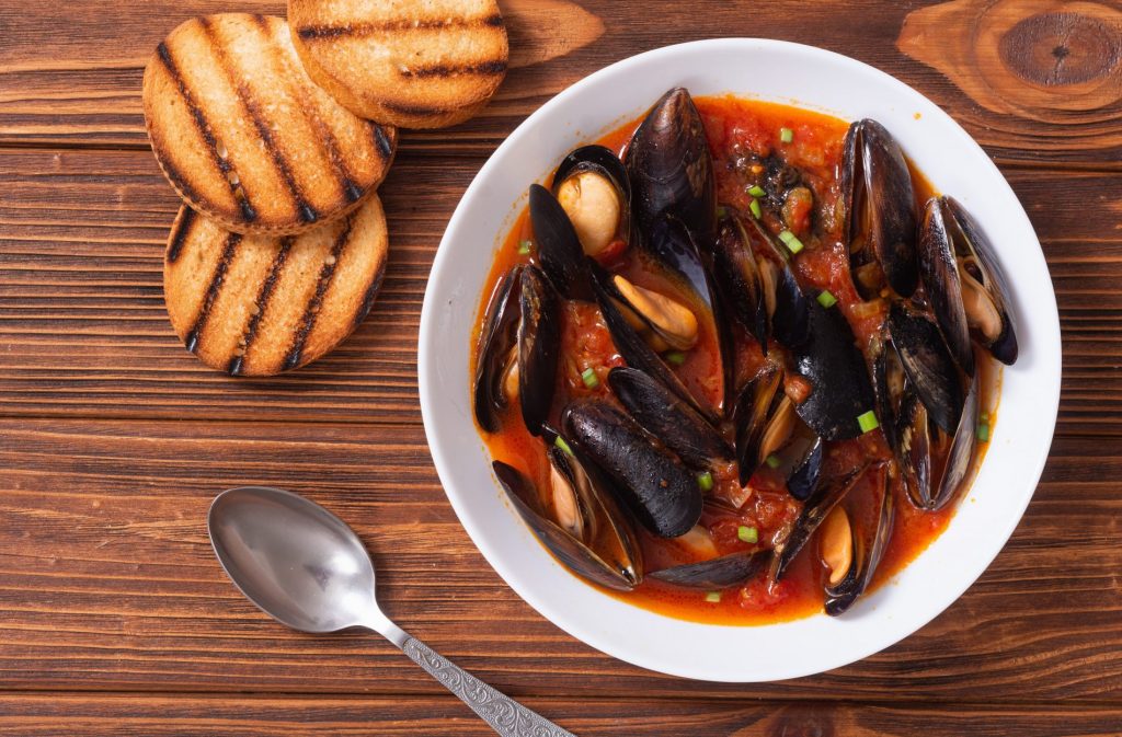 piatto bianco su un tavolo di legno con una zuppa di cozze, un cucchiaio e 3 fette di pane tostato