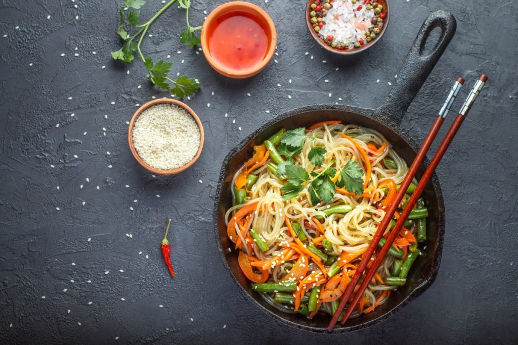 su base grigio scuro una padella con manico piena di spaghetti di riso con verdure, tre ciotole con salse varie, un paio di bacchette