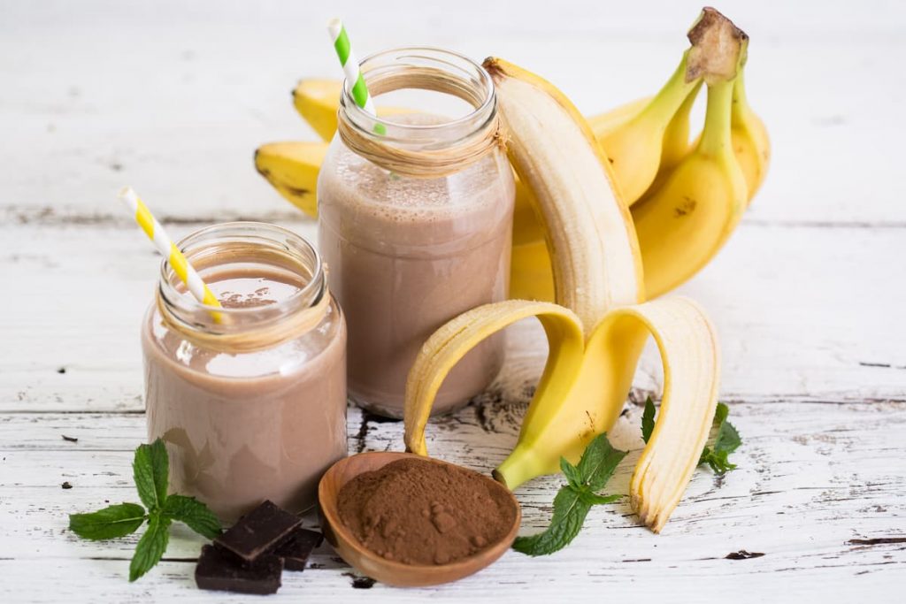 su sfondo bianco 2 bicchieri con frullati di banana e banane sbucciate e cacao amaro