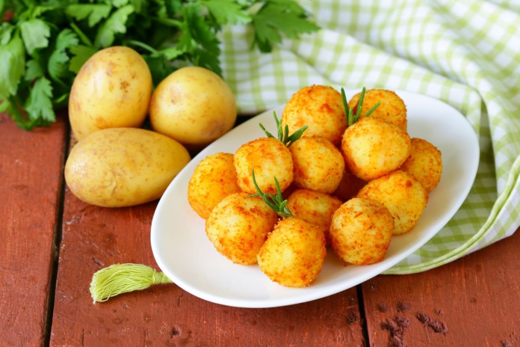 su tavolo di legno, piatto bianco con polpette di patate, accanto 3 patate e dietro prezzemolo e canovaccio verde a quadretti bianchi