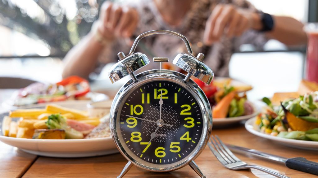 Donna che mangia su tavolo pieno di cibi con in primo piano un orologio per illustrare il digiuno intermittente